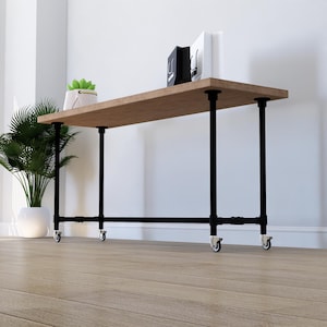 Puede incluir: Una consola de madera marrón con patas de metal negro y ruedas. La mesa tiene una parte superior rectangular y soporta una pequeña planta en maceta, bolsas de la compra y libros. La mesa está frente a una pared blanca.