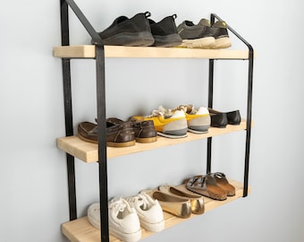 May include: A wall-mounted storage unit featuring three light-colored wooden shelves supported by two black metal frames. The shelves hold various styles of footwear, including sneakers, loafers, sandals, and flats, arranged across the three levels.