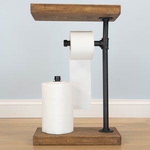May include: A rustic, freestanding toilet paper and paper towel holder. The holder features a dark stained wooden base and top shelf, connected by black metal pipes. It holds a roll of toilet paper and a roll of paper towels.