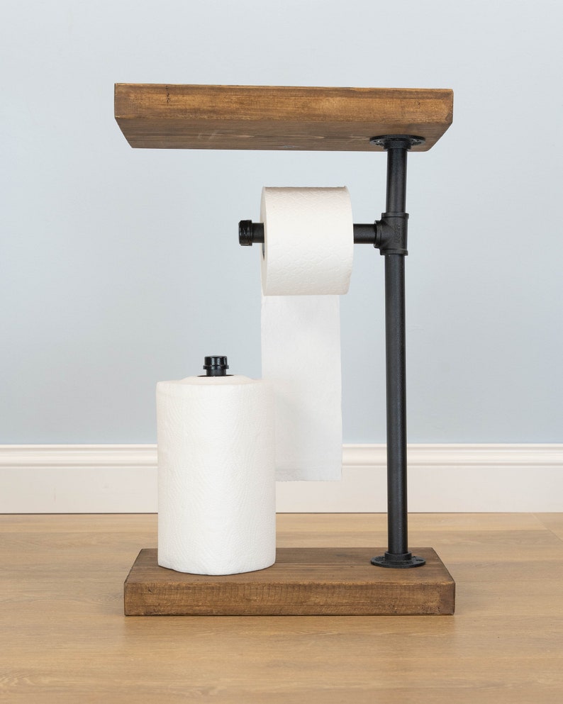 May include: A rustic, freestanding toilet paper and paper towel holder. The holder features a dark stained wooden base and top shelf, connected by black metal pipes. It holds a roll of toilet paper and a roll of paper towels.