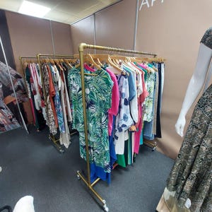 Pode incluir: Cabide com uma variedade de vestidos, tops e calças coloridas. As roupas são exibidas em cabides de madeira. O cabide é feito de metal dourado e tem rodas. Um manequim está no lado direito da imagem.