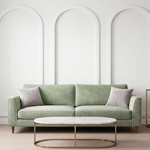 Puede incluir: Un sofá de terciopelo verde salvia con dos cojines gris claro, colocado detrás de una mesa de centro ovalada con tapa de mármol blanco y marco de metal dorado, junto a una mesa consola de metal dorado con un terrario de vidrio geométrico.