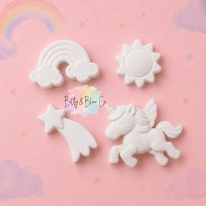 May include: Four white ceramic or clay shapes featuring a rainbow with clouds, a sun, a shooting star, and a winged unicorn. The items are arranged on a pink background with a rainbow and cloud graphic. The text Bitty & Blue Co. is centered.
