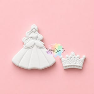 May include: A white cookie cutter shaped like a figure in a ballgown and a separate white cookie cutter shaped like a crown. The text Bitty & Blue Co. is printed in black script over a colorful watercolor splash between the two white shapes.