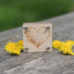 Könnte beinhalten: Eine Holzplakette mit einem Herzdesign und dem Text "Your name" eingraviert. Die Plakette ist von Löwenzahnblüten umgeben und hat vier silberne Metallfüße.