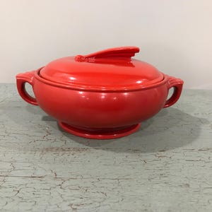 May include: A vibrant red ceramic serving bowl with a matching lid. The bowl has two handles and a pedestal base. The lid features a distinctive fin-shaped handle. The glossy finish enhances the bowl's bright color, making it a striking kitchenware piece.