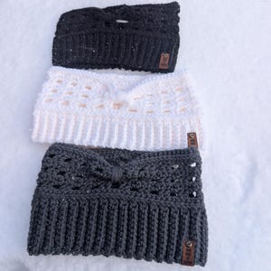 May include: Three crocheted headbands in black, white, and dark gray. Each headband features a ribbed bottom edge, a decorative knotted center, and a small brown rectangular tag with the text "HAND MADE BY" printed on the lower right corner.
