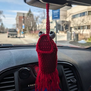 May include: A red crocheted lobster car air freshener with white googly eyes hanging from the rearview mirror of a car. The lobster has a fringe of red yarn.