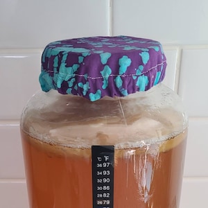 Op de afbeelding: Een heldere glazen pot met een paarse en turquoise stoffen hoes. De pot bevat een lichtbruine vloeistof. Een temperatuurstrip is aan de zijkant van de pot bevestigd.