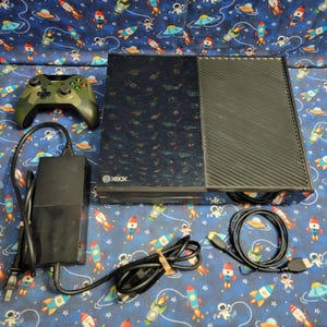 Puede incluir: Una consola de videojuegos Xbox negra con acabado brillante, un mando inalámbrico con patrón de camuflaje, un bloque de alimentación negro con cable y un cable HDMI negro. La consola presenta un panel lateral texturizado y el logo de Xbox.