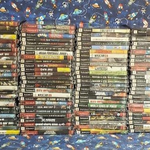 May include: A large collection of video game cases for the Playstation 2 console. The cases are stacked in rows and are mostly black with colorful titles and images. The games include titles like "Call of Duty", "Madden NFL", and "Tony Hawk's Pro Skater".
