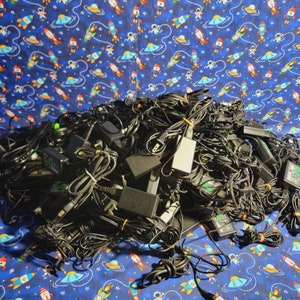 以下が含まれることがあります： 様々なプラグタイプを持つ、黒い電子電源アダプターと充電ケーブルが絡まり合った大きな山。電源ブロックとコードの集まりが積み重なっており、複数のコネクタと黒いプラスチック製の筐体が確認できます。