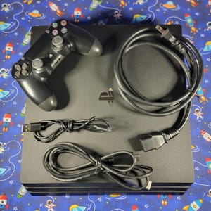 May include: A black PlayStation 4 games console with a matching black wireless controller, a black power lead, a black USB charging cable, and a black HDMI cable. The console features the PlayStation logo on the top surface of the square unit.
