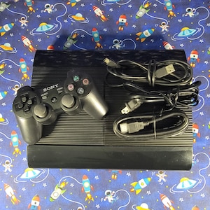 Puede incluir: Una consola de videojuegos Sony PlayStation 3 negra con un mando inalámbrico negro a juego y dos cables negros de alimentación y HDMI. La consola presenta una superficie superior estriada y un panel frontal liso, como un conjunto completo.
