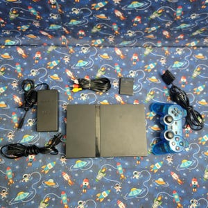 Op de afbeelding: Een zwarte PlayStation 2-console met een blauwe doorschijnende controller, voedingsadapter, kabels en een geheugenkaart. De console is omgeven door accessoires op een blauwe stof met raketten en astronauten.