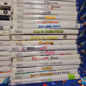 K&ouml;nnte beinhalten: Ein Stapel von 15 Videospiel-H&uuml;llen f&uuml;r die Nintendo Wii-Konsole. Die H&uuml;llen sind wei&szlig; mit bunten Spieltiteln und Logos. Die Titel umfassen "Wii Music", "Big Beach Sports", "Spore Hero", "Ben 10 Alien Force", "Kung Fu Panda", "Punch-Out!!", "Madden NFL 07", "Wii Party", "Rec Room Games", "Cyberbike Cycling Sports", "Thrillville Off the Rails", "Animal Crossing City Folk" und "Wii Fit".