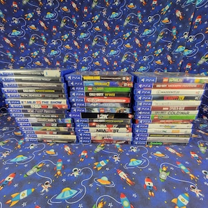 Może przedstawiać: Trzy pionowe stosy pudełek z grami wideo na PlayStation 4 ułożone na niebieskiej tkaninie z powtarzającym się wzorem kolorowych rysunkowych astronautów, rakiet i latających spodków. Tytuły gier są widoczne na grzbietach pudełek.