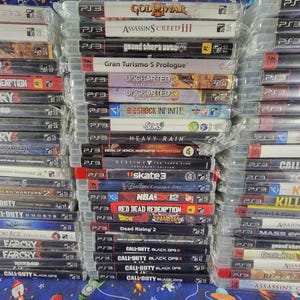May include: A large collection of PlayStation 3 video game cases stacked in three vertical columns. The spines display various titles including God of War, Assassin's Creed, Grand Theft Auto, Uncharted 3, Bioshock Infinite, The Sims 3, and Call of Duty.