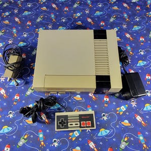 May include: A beige and black Nintendo Entertainment System game console with a rectangular controller, a black power adapter, and a grey AV cable. The console features a front-loading cartridge slot and a black panel on the right side.