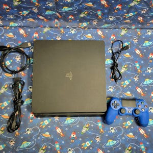 May include: A black games console with a blue controller and connecting cables. The controller has multiple buttons and two joysticks. The console has the PlayStation logo on the top. The background is a blue fabric with a pattern of rockets, astronauts, and UFOs.