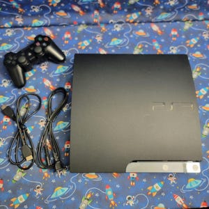 Könnte beinhalten: Eine schwarze PlayStation 3-Konsole mit passendem Controller und Verbindungskabeln. Die Konsole hat eine silberne Zierleiste und das PS3-Logo. Der Controller ist schwarz mit mehreren Tasten und Joysticks. Der Hintergrund ist ein blauer Stoff mit Weltraum-Motiv.