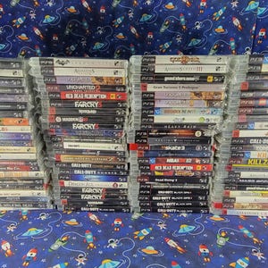 May include: A large collection of PlayStation 3 video game cases stacked in four vertical columns. Each case features a distinct spine with game titles and logos, all encased in clear plastic wrap, showcasing a variety of titles for the console.