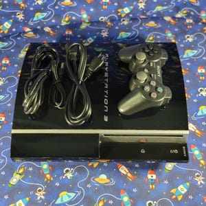 May include: A black Sony PlayStation 3 game console with a glossy finish, featuring the text PLAYSTATION 3 on the top surface. The console includes a black wireless controller and two black power and connection cables resting on the unit.