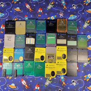 May include: A collection of thirty-one rectangular PlayStation memory cards in various colors including black, yellow, green, blue, gray, and translucent orange. Each card features the PlayStation logo and text indicating 8MB storage capacity.