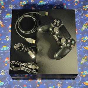May include: A black rectangular game console with a matching black wireless controller featuring dual analog sticks, a directional pad, and four action buttons. Included are a black HDMI cable, a USB charging cable, and a power cord.