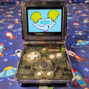 May include: A transparent dark grey Game Boy Advance SP handheld gaming console with a clear shell revealing internal components. The screen displays a cartoon character with blonde pigtails and large blue eyes. It features white buttons.