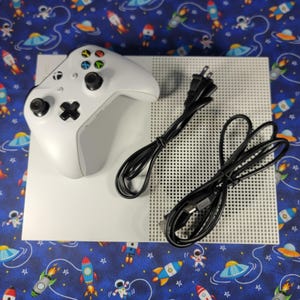 Puede incluir: Una consola de videojuegos Xbox One S blanca con un mando inalámbrico blanco a juego que presenta joysticks negros y botones de colores, acompañada de un cable de alimentación negro y un cable HDMI negro, todo sobre una superficie plana.