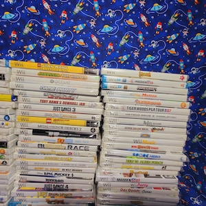 K&ouml;nnte beinhalten: Ein Stapel von &uuml;ber 30 wei&szlig;en Wii-Videospielh&uuml;llen mit bunten Spieltiteln und Logos. Die H&uuml;llen sind auf einem blauen Hintergrund mit einem Muster aus Sternen, Raketen und Raumschiffen gestapelt.