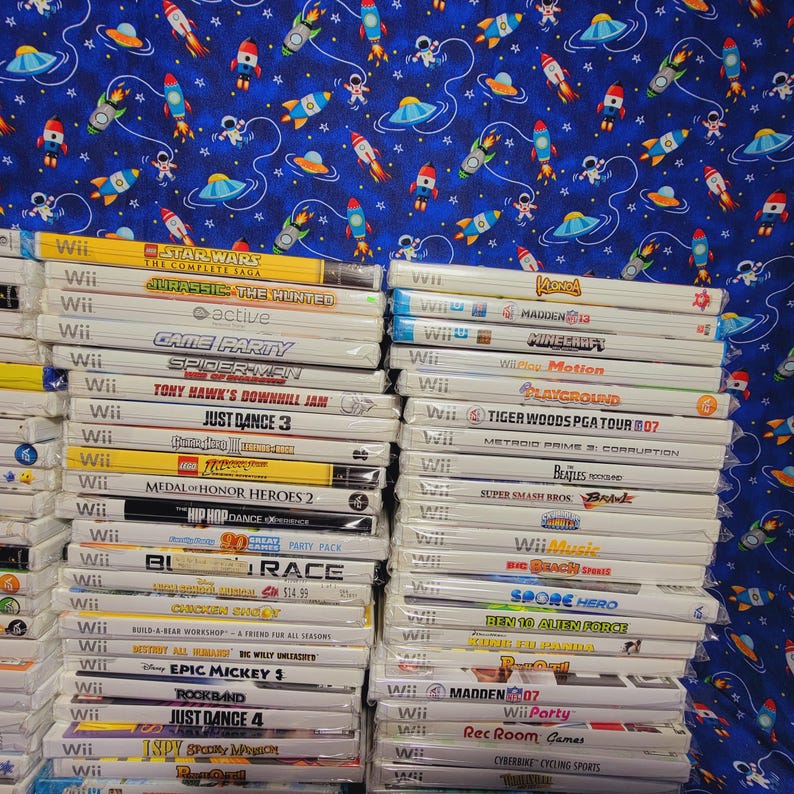 K&ouml;nnte beinhalten: Ein Stapel von &uuml;ber 30 wei&szlig;en Wii-Videospielh&uuml;llen mit bunten Spieltiteln und Logos. Die H&uuml;llen sind auf einem blauen Hintergrund mit einem Muster aus Sternen, Raketen und Raumschiffen gestapelt.