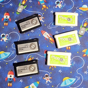 Könnte beinhalten: Vier Nintendo DS Rumble Pak-Kartuschen und drei Motion Pak-Kartuschen. Die Rumble Pak-Kartuschen sind schwarz mit einem silbernen Nintendo DS-Logo und dem Text "Nintendo DS Rumble Pak" darauf gedruckt. Die Motion Pak-Kartuschen sind weiß mit einem grünen Nintendo-Logo und dem Text "Motion Pak" darauf gedruckt.