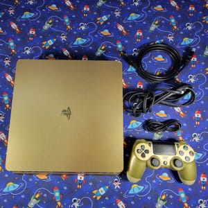 May include: A gold-coloured game console with the PlayStation logo on top, accompanied by a matching gold wireless controller and three black connection cables. The console has a square, flat design with rounded corners and a smooth metallic finish.