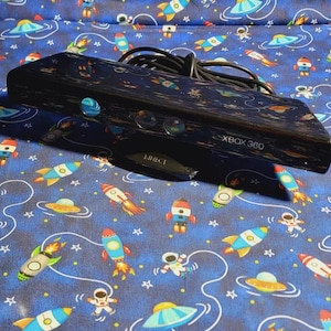 Könnte beinhalten: Ein schwarzer Xbox 360 Kinect-Sensor mit Weltraum-Design. Der Sensor hat eine dunkelblaue Oberfläche mit Planeten und Sternen. Die Texte "XBOX 360" und "KINECT" sind sichtbar. Der Sensor liegt auf einem blauen Stoff mit Raketen, Astronauten und UFOs.