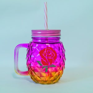 K&ouml;nnte beinhalten: Ein farbenfrohes Mason-Glas mit Farbverlauf von Gelb nach Pink und einem Rosendesign. Es hat einen pinkfarbenen Deckel und Griff sowie einen rosa-wei&szlig; gestreiften Strohhalm. Geeignet f&uuml;r kalte Getr&auml;nke.