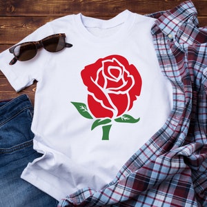 K&ouml;nnte beinhalten: Wei&szlig;es T-Shirt mit einer gro&szlig;en roten Rosengrafik mit gr&uuml;nen Bl&auml;ttern und Stiel. Das Shirt wird mit Sonnenbrille, kariertem Hemd und Jeans auf einer Holzoberfl&auml;che pr&auml;sentiert. Ein l&auml;ssiger Artikel mit floralem Thema.
