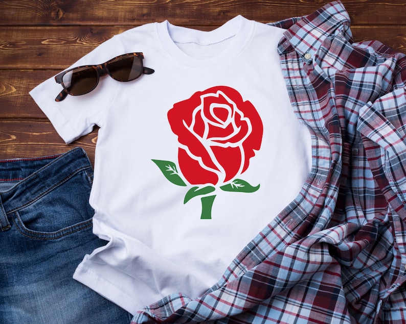 K&ouml;nnte beinhalten: Wei&szlig;es T-Shirt mit einer gro&szlig;en roten Rosengrafik mit gr&uuml;nen Bl&auml;ttern und Stiel. Das Shirt wird mit Sonnenbrille, kariertem Hemd und Jeans auf einer Holzoberfl&auml;che pr&auml;sentiert. Ein l&auml;ssiger Artikel mit floralem Thema.