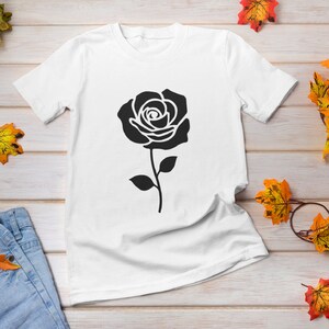 May include: White t-shirt featuring a large black rose graphic. The rose is centered on the shirt and has detailed petals and a stem with leaves. The shirt is displayed on a light wood surface with autumn leaves.