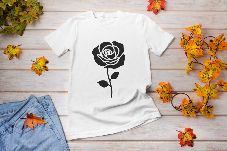 May include: White t-shirt featuring a large black rose graphic. The rose is centered on the shirt and has detailed petals and a stem with leaves. The shirt is displayed on a light wood surface with autumn leaves.