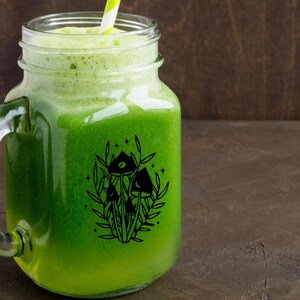May include: A mason jar filled with green juice and a black and white illustration of mushrooms and stars on the side.