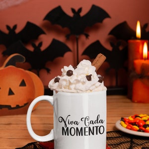 Op de afbeelding: Een witte mok met de tekst "Viva Cada Momento" is gevuld met slagroom, chocolade chips en een kaneelstokje. De mok staat op een rode onderzetter, met Halloween-decoraties op de achtergrond.