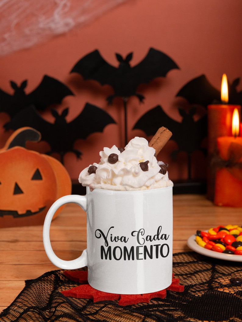Op de afbeelding: Een witte mok met de tekst "Viva Cada Momento" is gevuld met slagroom, chocolade chips en een kaneelstokje. De mok staat op een rode onderzetter, met Halloween-decoraties op de achtergrond.