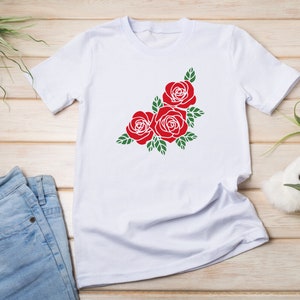 K&ouml;nnte beinhalten: Wei&szlig;es T-Shirt mit einem roten Rosenmuster und gr&uuml;nen Bl&auml;ttern. Die Rosen sind in einer Ecke des Shirts angeordnet. Das T-Shirt wird auf einer Holzoberfl&auml;che mit einer Jeans pr&auml;sentiert.