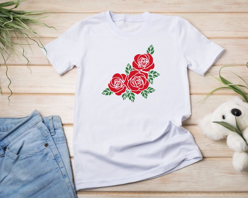 K&ouml;nnte beinhalten: Wei&szlig;es T-Shirt mit einem roten Rosenmuster und gr&uuml;nen Bl&auml;ttern. Die Rosen sind in einer Ecke des Shirts angeordnet. Das T-Shirt wird auf einer Holzoberfl&auml;che mit einer Jeans pr&auml;sentiert.