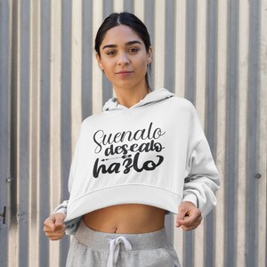 Op de afbeelding: Witte cropped hoodie met de tekst "Suenalo, desealo, hazlo" in zwart script. De hoodie heeft een capuchon met trekkoord en lange mouwen. De persoon draagt een grijze joggingbroek.