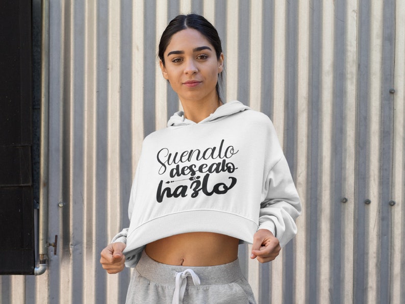 Op de afbeelding: Witte cropped hoodie met de tekst "Suenalo, desealo, hazlo" in zwart script. De hoodie heeft een capuchon met trekkoord en lange mouwen. De persoon draagt een grijze joggingbroek.