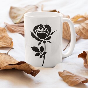 May include: White ceramic mug featuring a black rose illustration. The rose design is centered on the mug, with a stem and leaves. The mug is surrounded by dried brown leaves, creating a natural aesthetic.