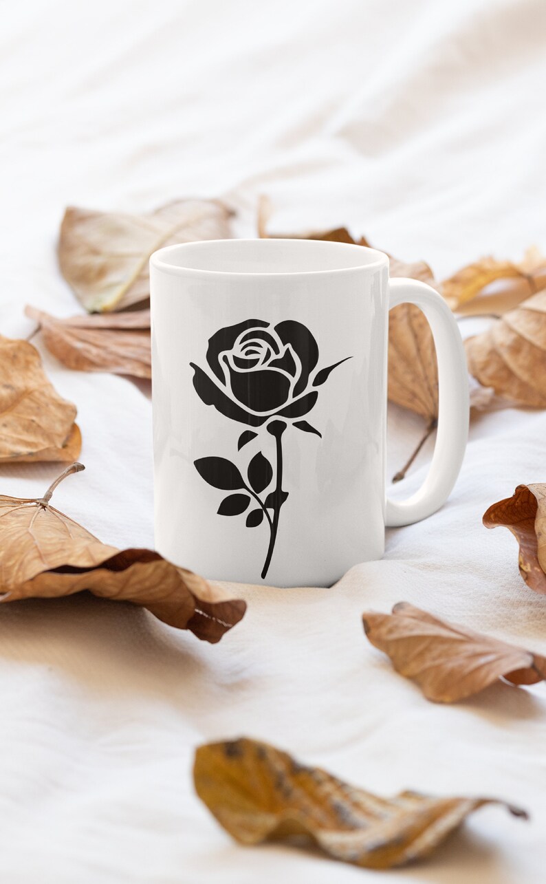May include: White ceramic mug featuring a black rose illustration. The rose design is centered on the mug, with a stem and leaves. The mug is surrounded by dried brown leaves, creating a natural aesthetic.